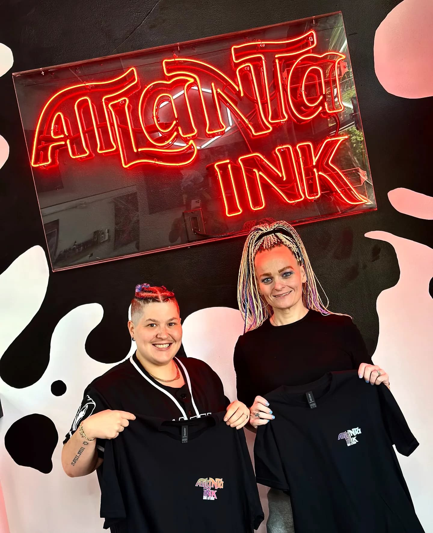 Two guests posing under the ATLANTA INK neon sign, holding branded T-shirts