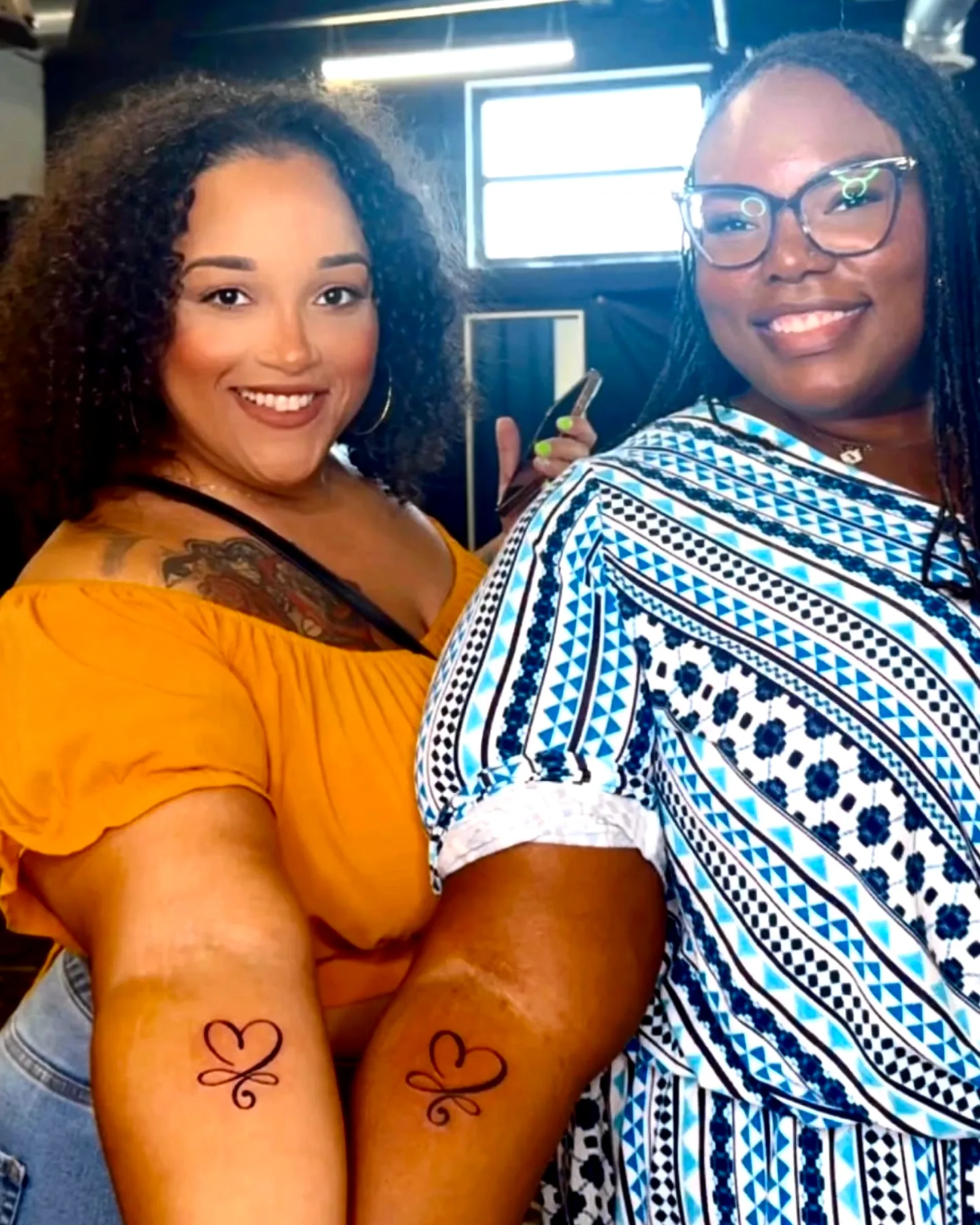 Two smiling clients holding up matching heart tattoos on their forearms